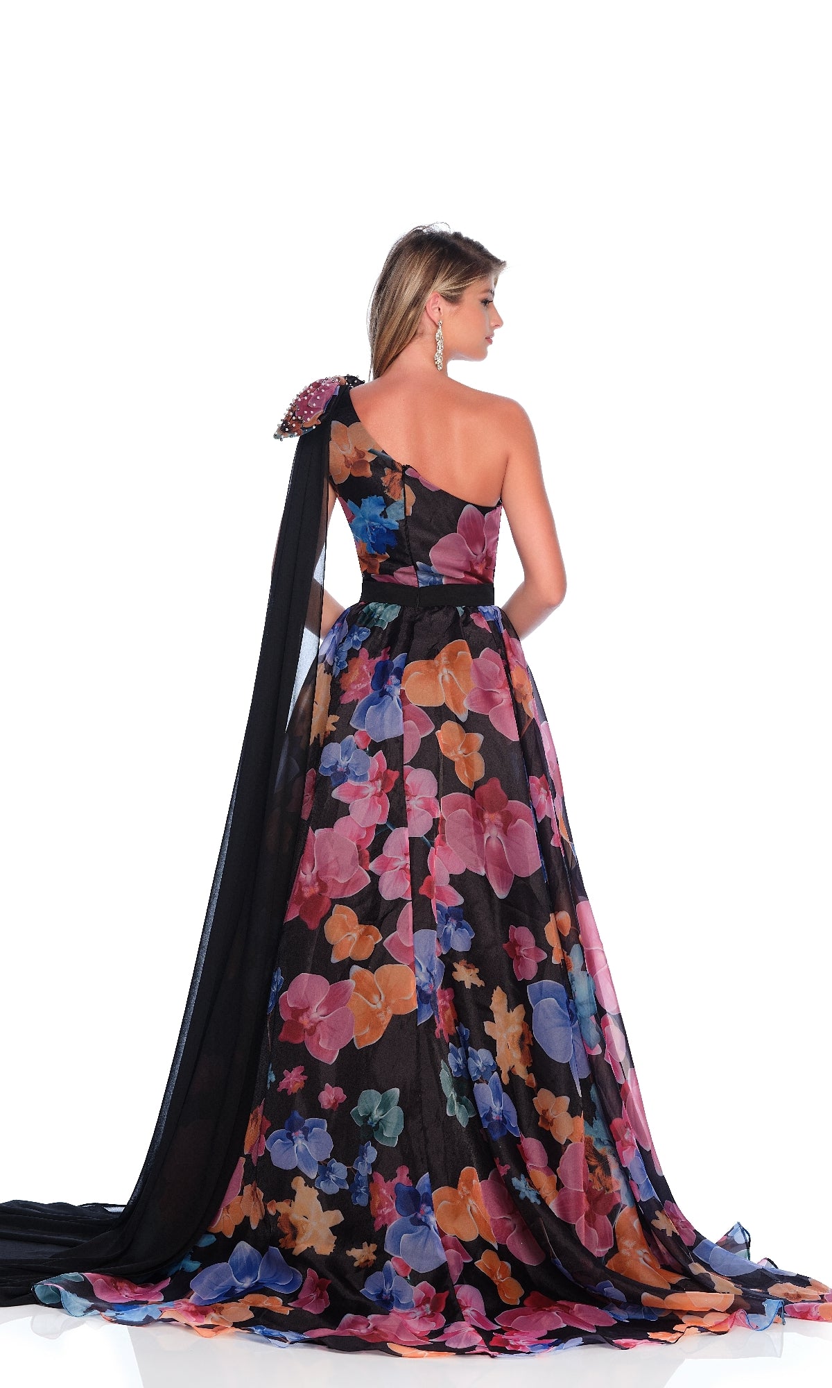 A woman faces away, wearing the Dave & Johnny 12157 Floral Print Prom Dress with a one-shoulder neckline, floor-length A-line skirt, and a long black drape over one shoulder.